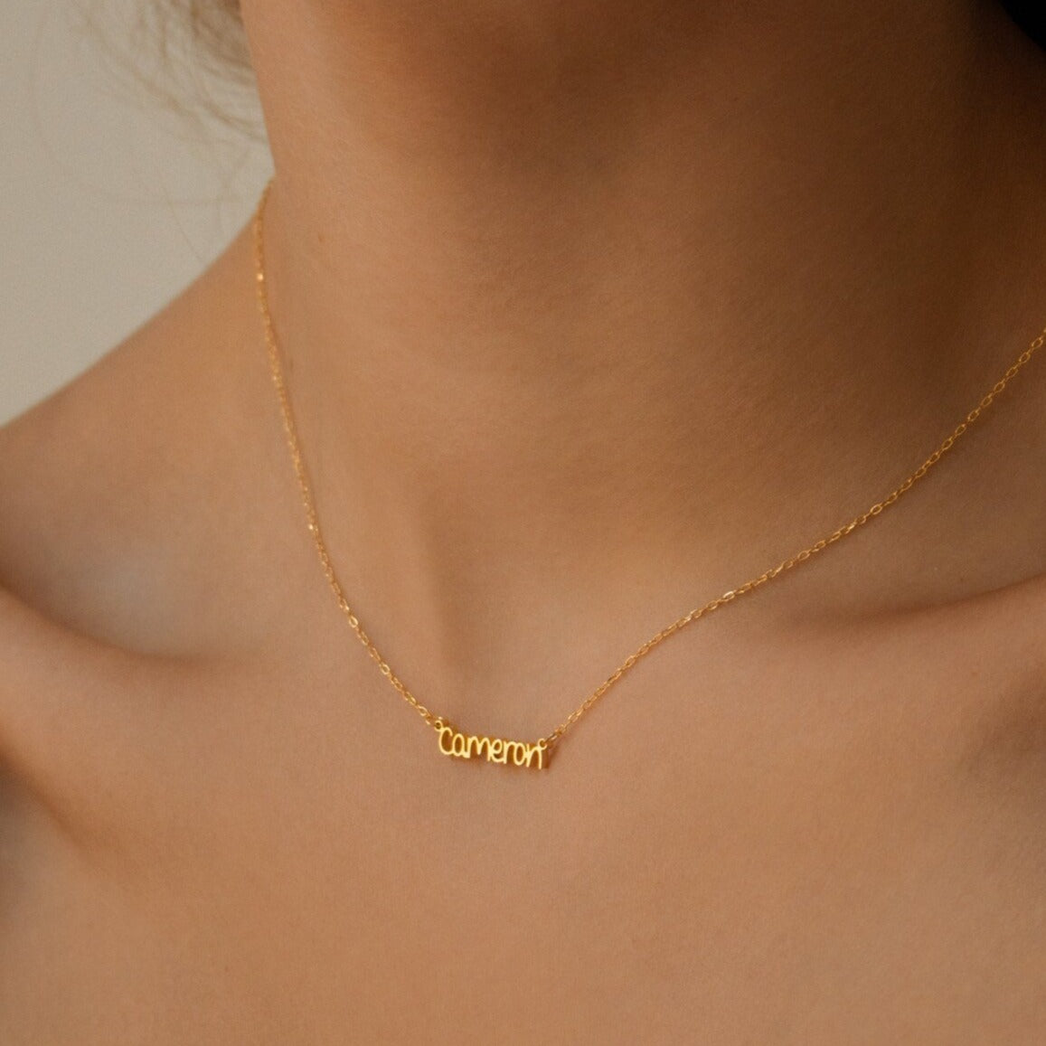 The Tiny Preppy Name Necklace, shown worn by a woman from the shoulders up with "Cameron" in a youthful font, is fully customizable with any name of your choice.