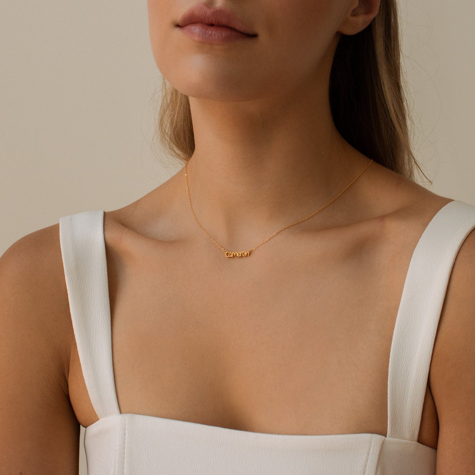 A woman in a white top wears the Tiny Preppy Name Necklace, a gold necklace with a small customizable nameplate charm in a youthful font.