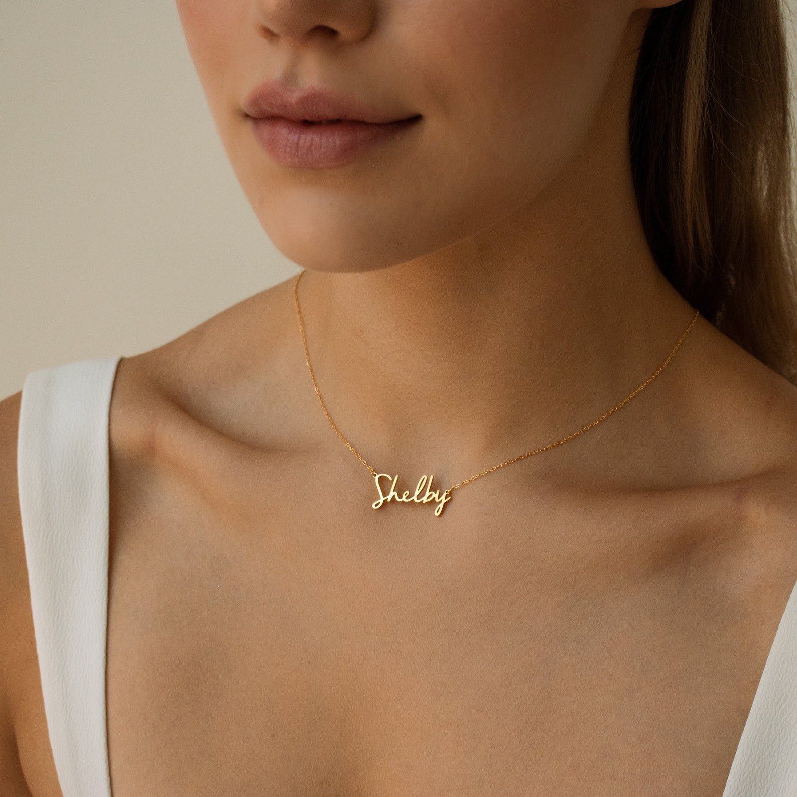 Woman wearing the Nella Name Necklace, a custom gold nameplate in cursive script with "Shelby," paired with a white top.