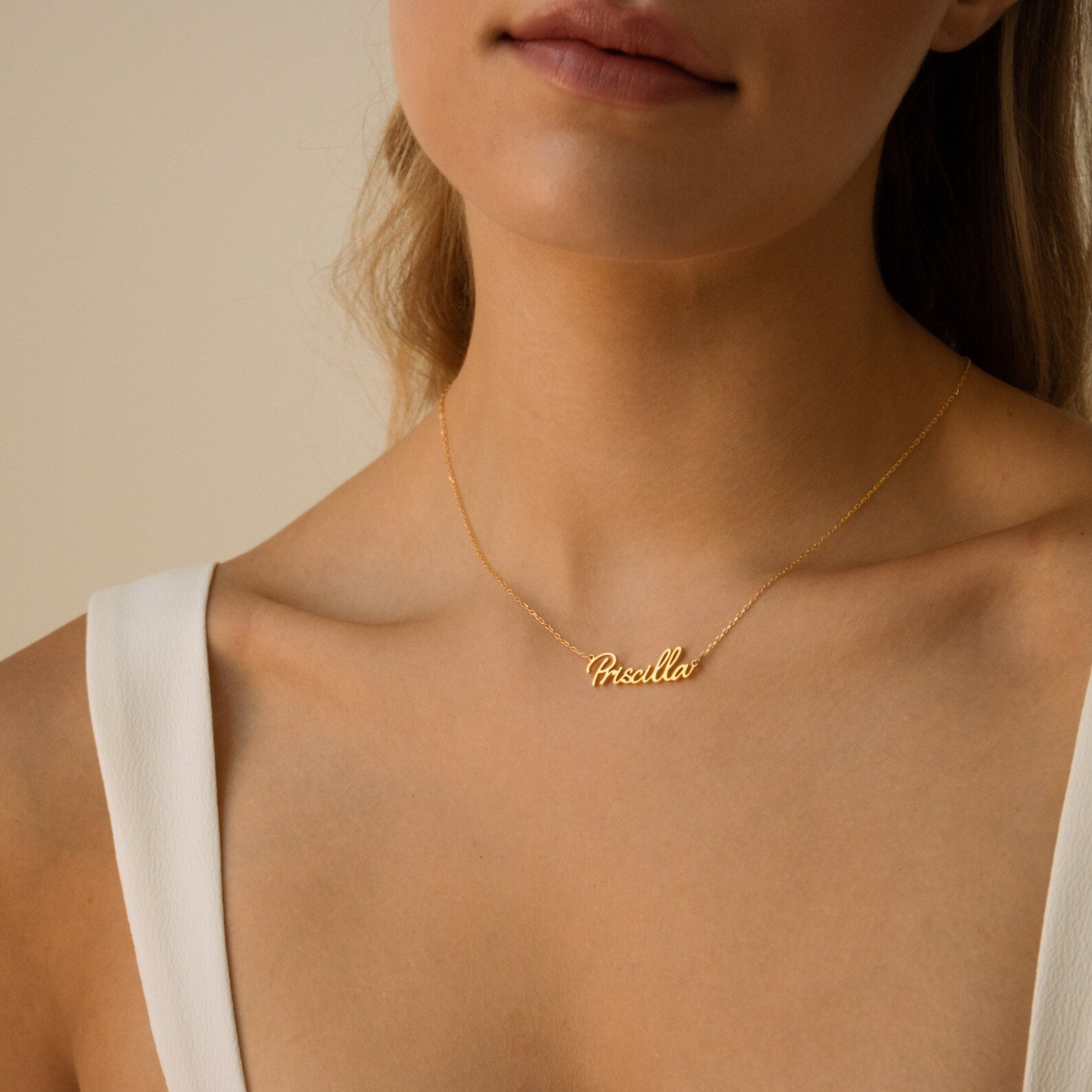 A woman elegantly styles the Grace Name Necklace in gold, featuring the name Priscilla in cursive, paired with a chic white top.