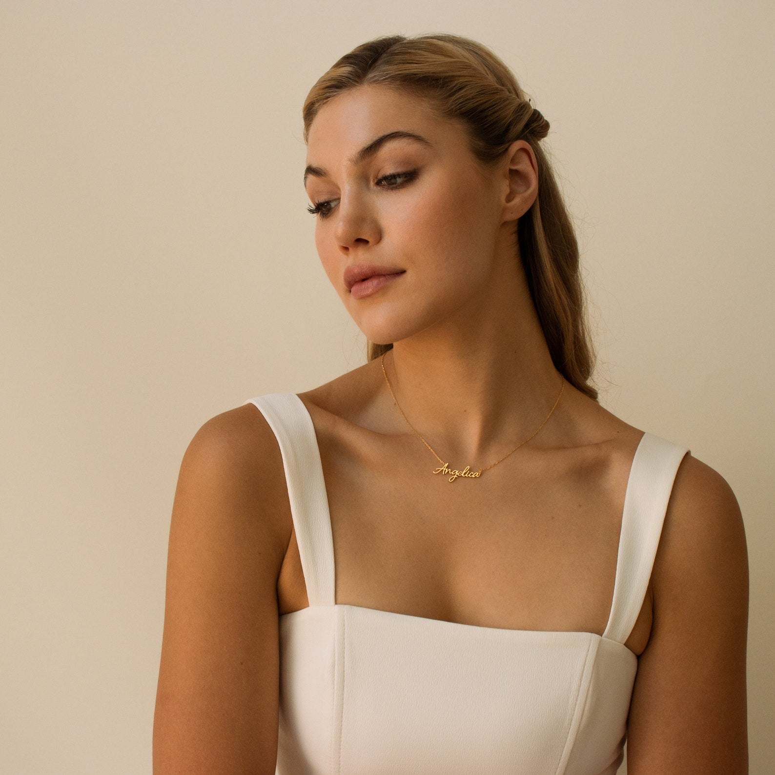 A woman in a white dress looks down, wearing the Grace Name Necklace—a personalized gold necklace featuring her name pendant—set against a beige background.