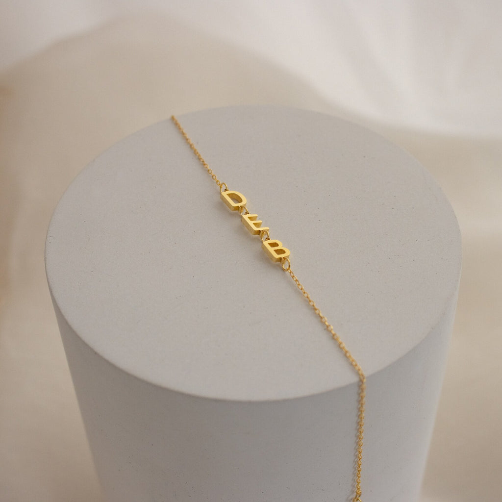 The Custom Letter Bracelet features gold QUEEN lettering on a gold chain, displayed on a round light gray pedestal. Ideal for fans of custom letter bracelet styles and unique personalized name jewelry.