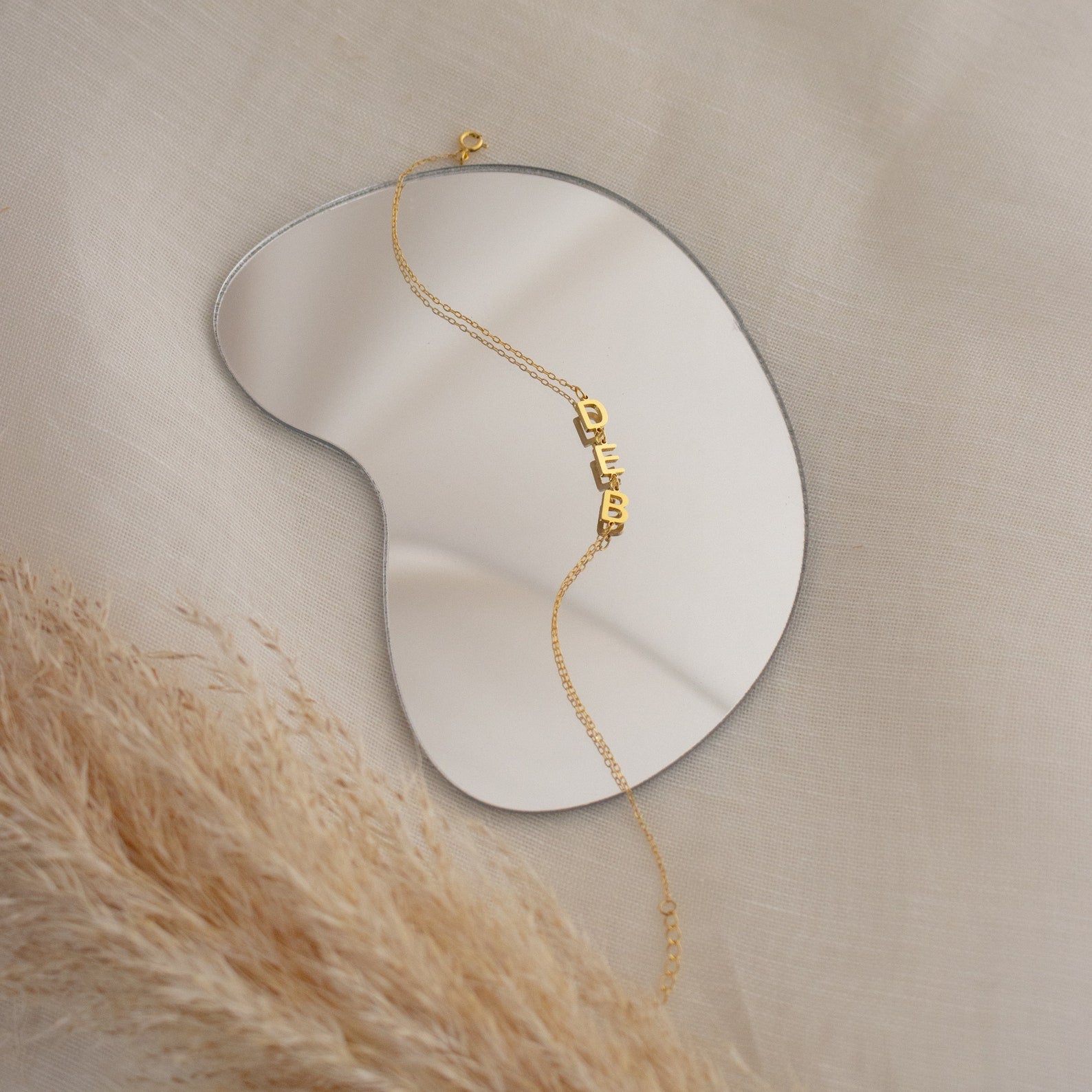 Custom Letter Bracelet displayed on a kidney-shaped mirror with beige fabric and pampas grass—ideal inspiration for personalized name jewelry and charm bracelets.