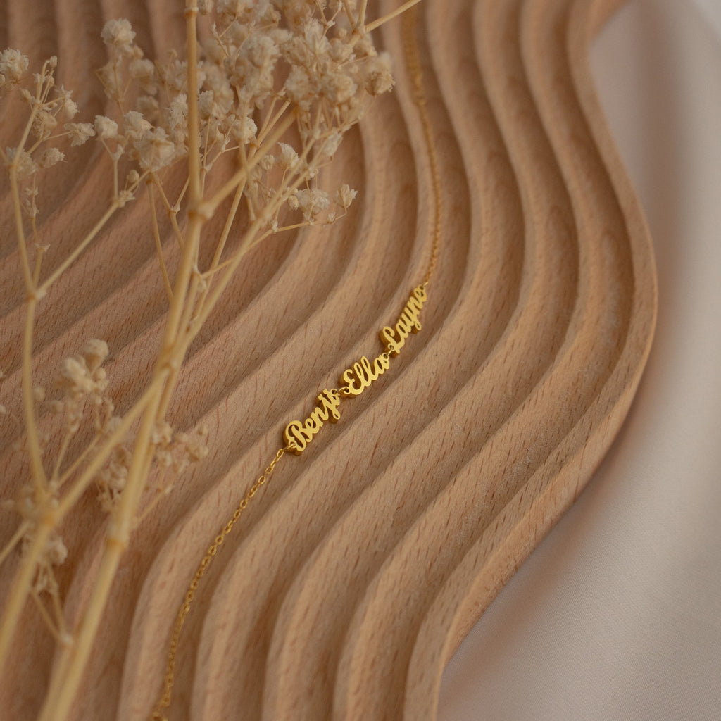 Gold name necklace on a wavy wooden surface with dried flowers and soft fabric in the background, perfect for pairing with dainty jewelry like the London Multiple Name Bracelet.
