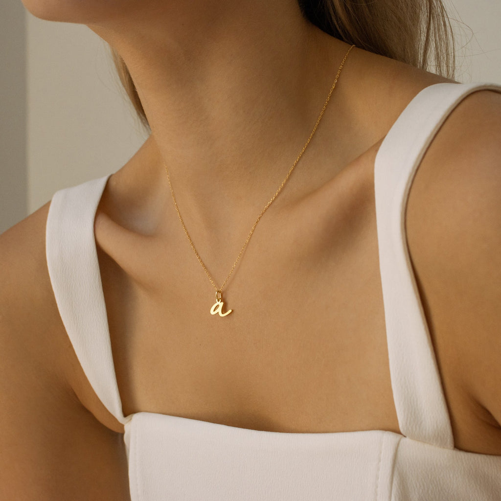 A woman wears a white sleeveless top and the Nella Letter Necklace featuring a gold lowercase "a" pendant.
