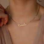 The Nella Name Necklace in Box Chain, featuring "Natalie," rests on a neck as a hand gently touches the chain, styled with a beige top.
