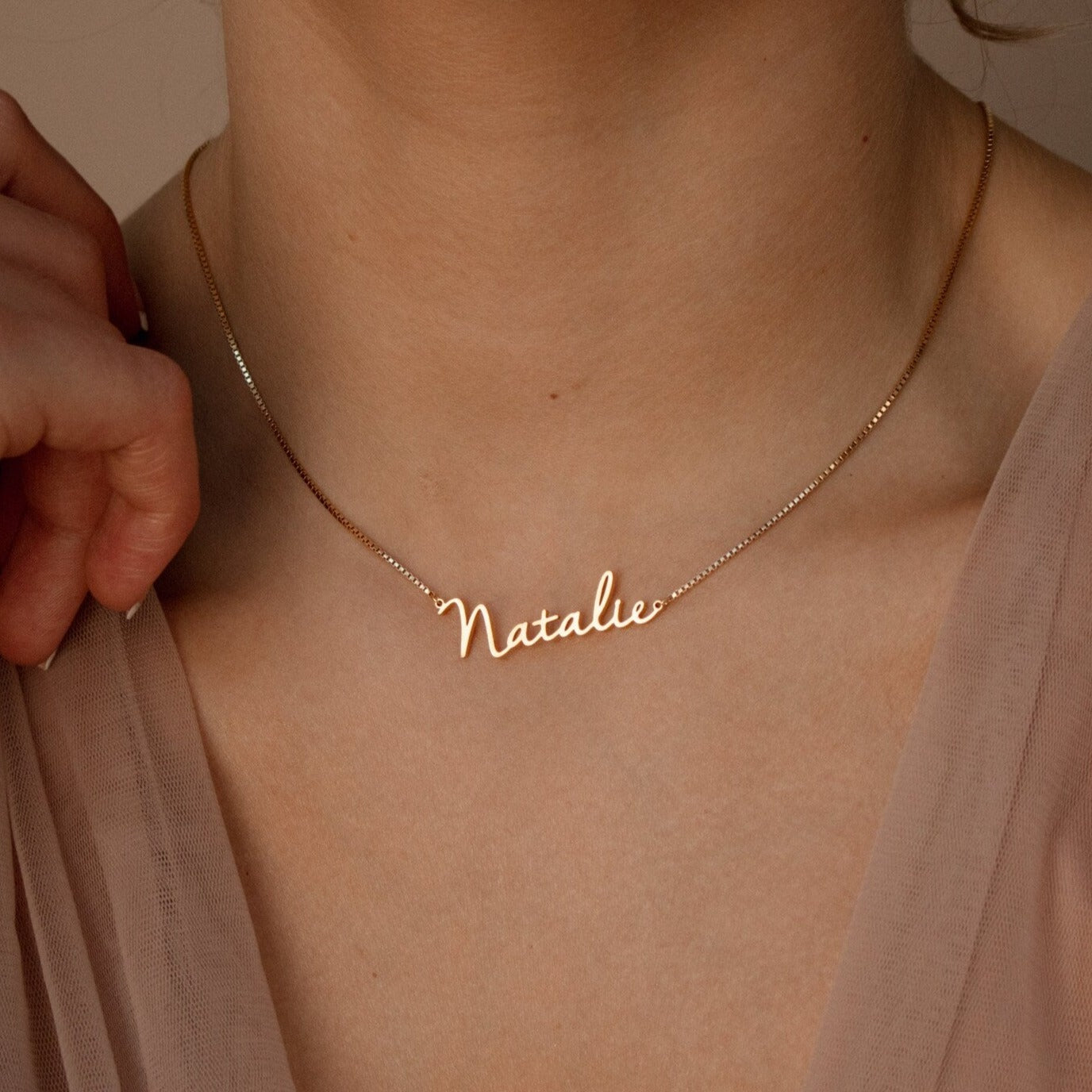 The Nella Name Necklace in Box Chain, featuring "Natalie," rests on a neck as a hand gently touches the chain, styled with a beige top.