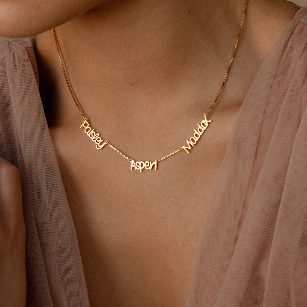 The Preppy Multiple Name Necklace in Box Chain, shown in gold with the names Paisley, Aspen, and Maddox in a youthful font, is styled here with a sheer top.