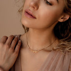 A woman wears the Callie Box Chain Name Necklace in 18K Gold, featuring a customizable charm on a delicate chain, as she touches her collarbone and looks slightly down.