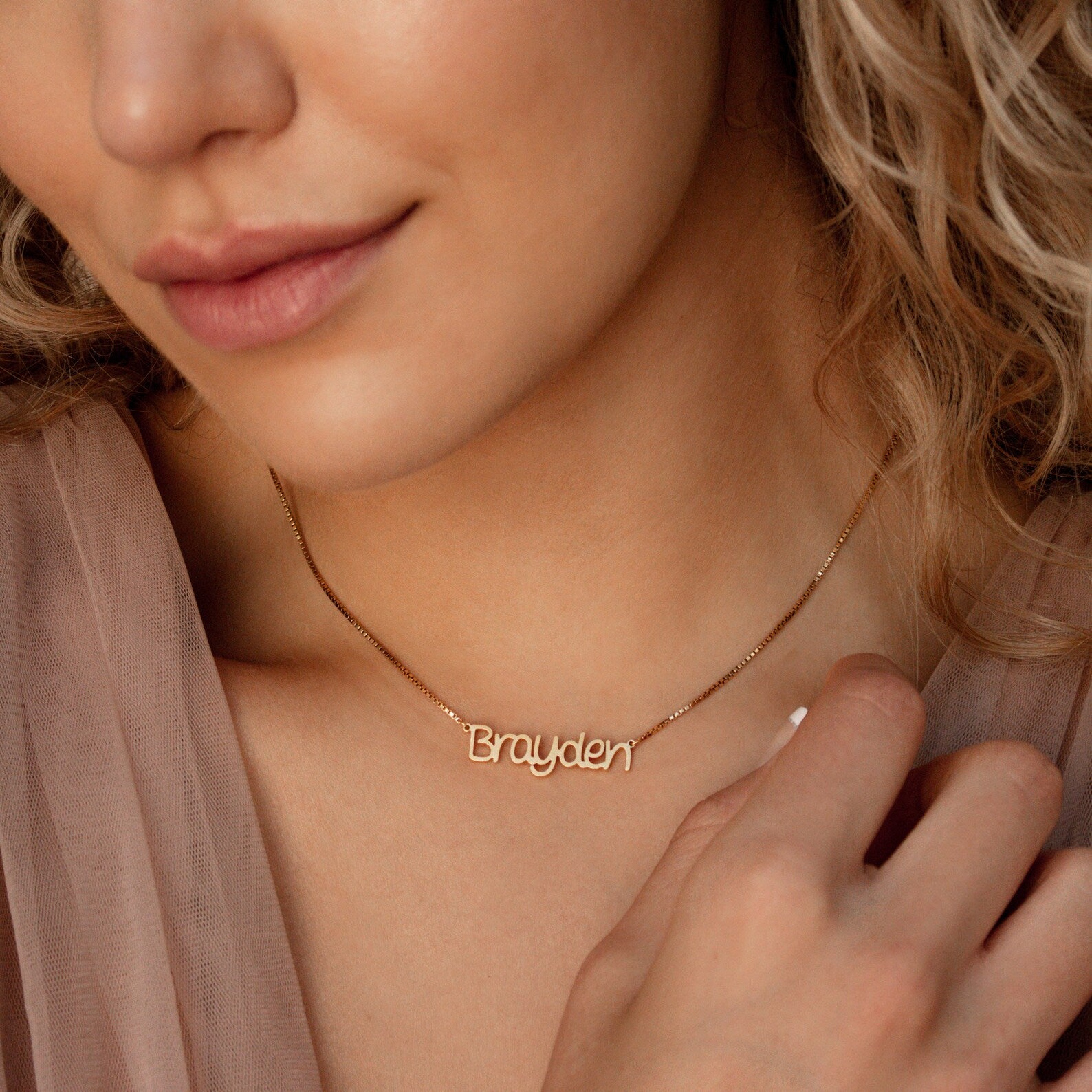A woman wears the Nella Name Necklace in Box Chain, featuring the name Brayden in cursive letters, as she gently touches the elegant chain.