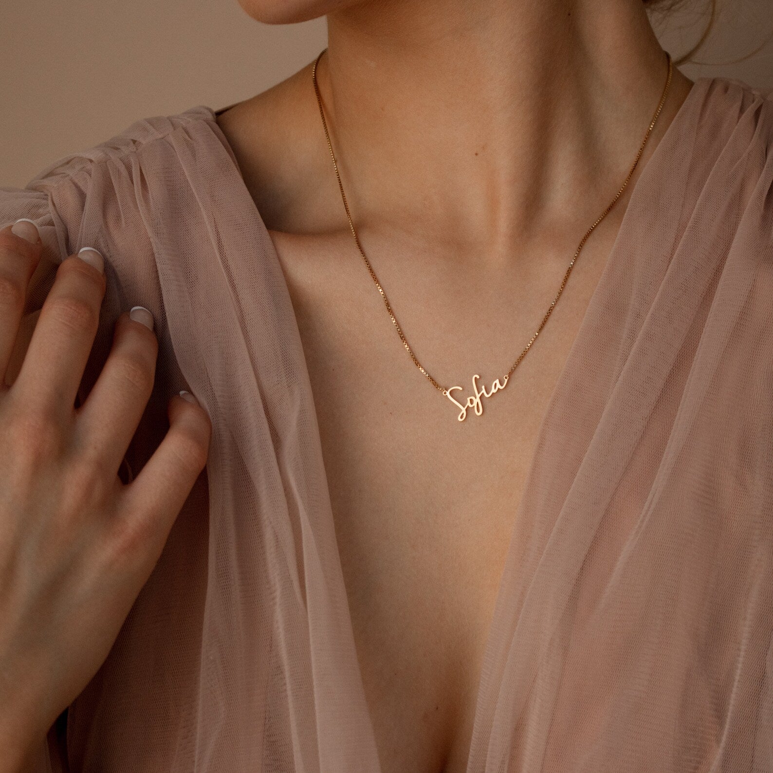 A woman wears a sheer pink blouse and the Nella Name Necklace in Box Chain featuring a gold "Sofia" pendant.