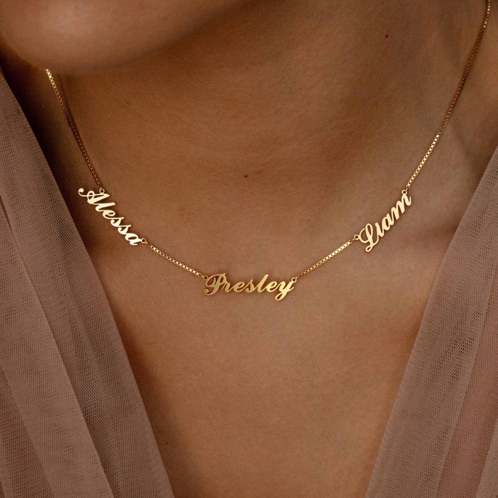 Close-up of a gold Carrie Multiple Name Necklace in Box Chain, featuring the names Alessa, Presley, and Gianni in cursive on a person's neck.