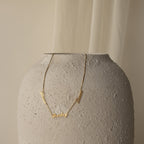 Carrie Multiple Name Necklace in Box Chain draped over a textured, light gray ceramic vase with a soft beige background, highlighting elegant personalized jewelry.