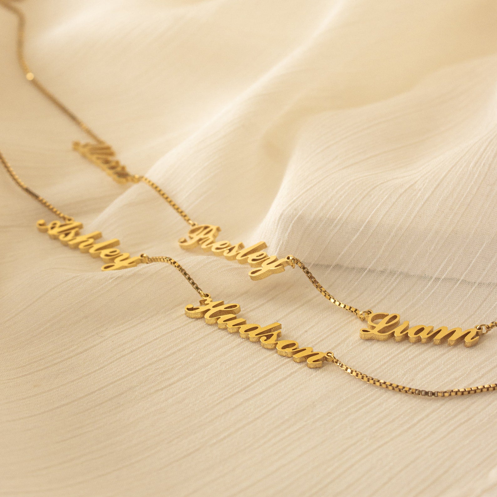 The Carrie Multiple Name Necklace in Box Chain, featuring personalized names on a delicate chain, is displayed on a light, textured fabric background—perfect for fans of custom jewelry.