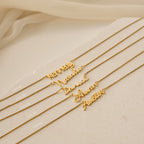 Five Callie Box Chain Name Necklaces in 18K Gold are displayed in various fonts on a light beige surface, with soft fabric in the background.