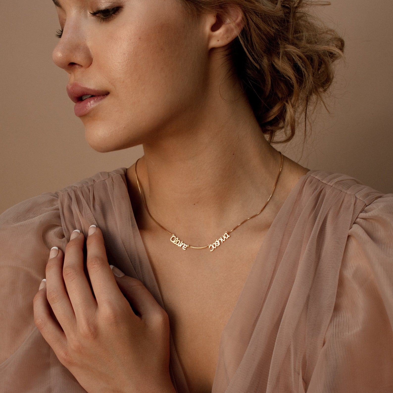 Woman wearing a sheer beige blouse and a personalized Preppy Multiple Name Necklace in Box Chain, featuring a youth-inspired font, posing with her hand near her neck.