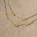 Showcasing the Preppy Multiple Name Necklace in Box Chain: five gold necklaces with personalized names—Naomi, Evelyn, Kylie, Taylor, and Jessica—in a chic youth-inspired font on a beige stone surface.
