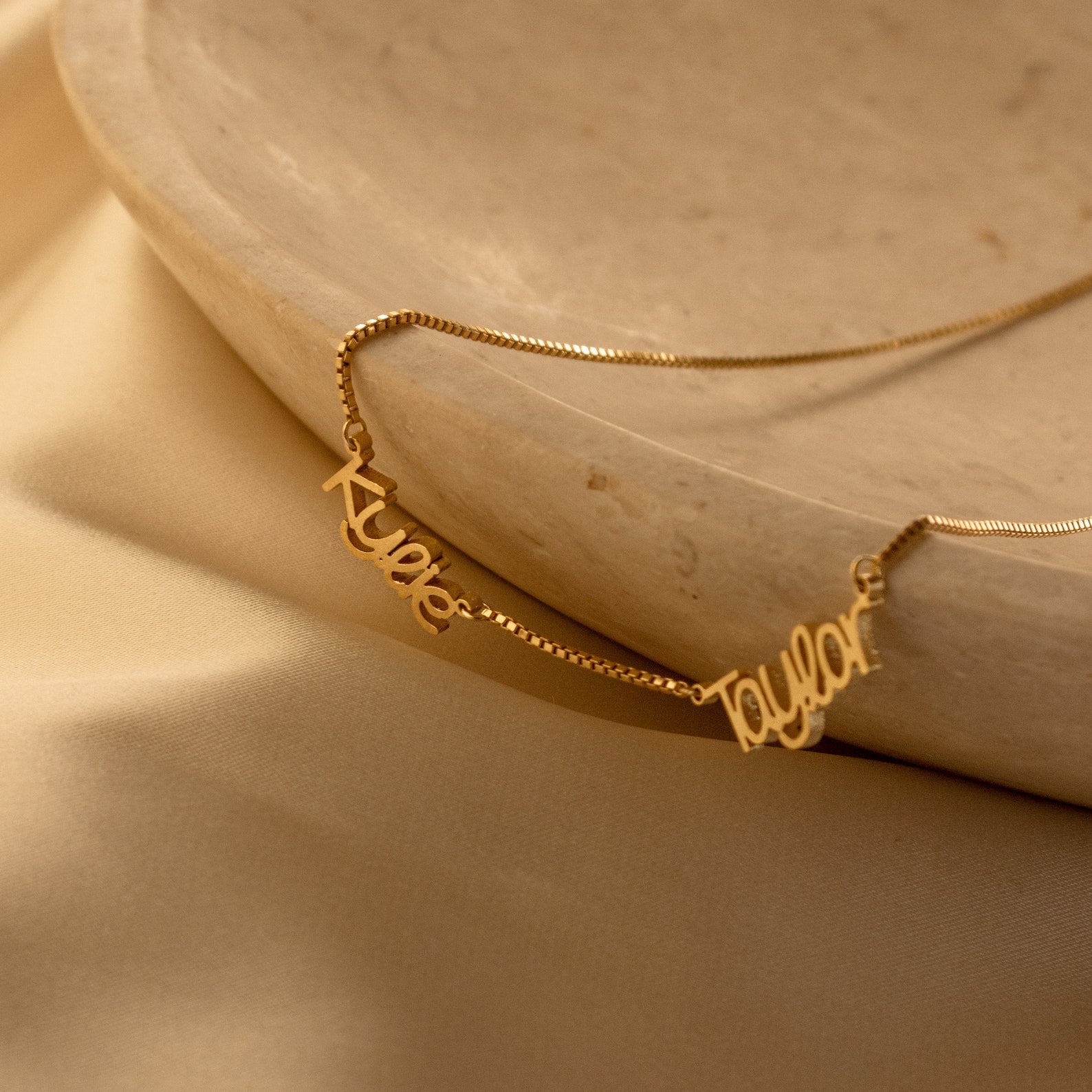 The Preppy Multiple Name Necklace in Box Chain features names like "Kyle" and "Taylor" in a youthful font, displayed on a beige stone dish with a satin background—a stylish, personalized jewelry piece.