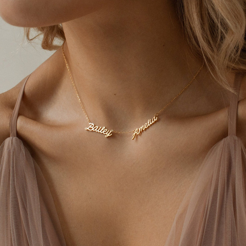 Close-up of a woman wearing the Paris Multiple Name Necklace in gold with Bailey and Amelia pendants, styled with taupe fabric.