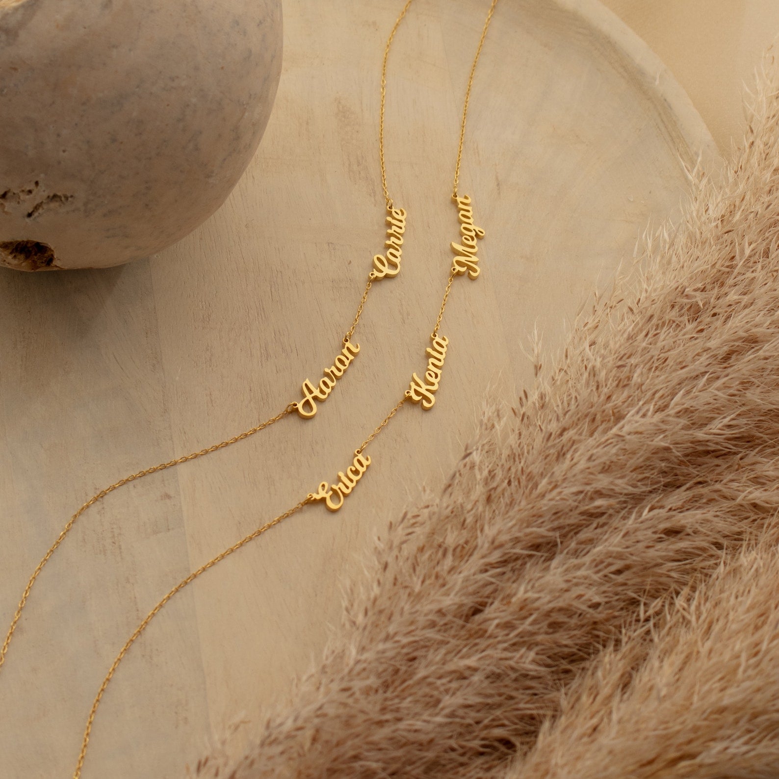 Four Mellow Multiple Name Necklaces in gold, customized with names Anna, Katie, Megan, and Erica, are displayed on wood with pampas grass—thoughtful personalized jewelry and a perfect gift for mom.