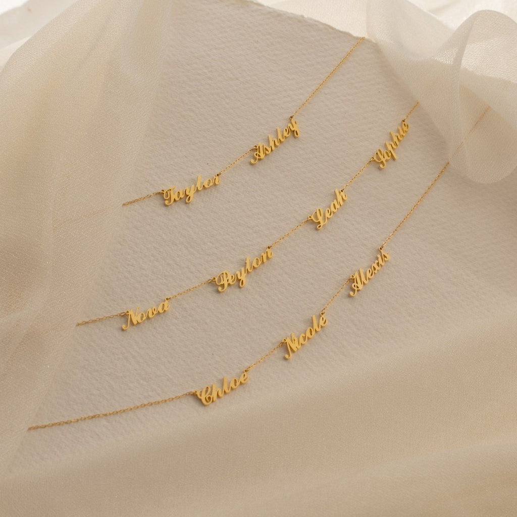The Carrie Multiple Name Necklace is displayed on textured cream fabric with sheer beige fabric draped above.