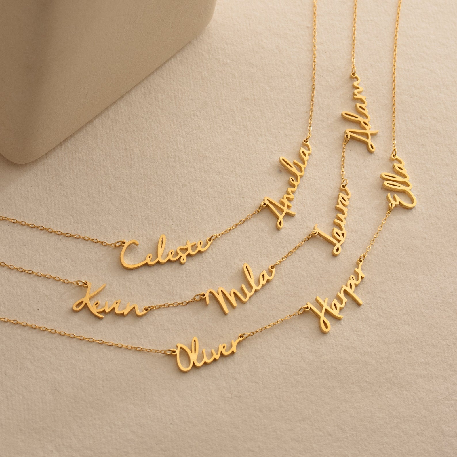 Five gold "Nella Multiple Name Necklaces" with cursive name pendants—Celeste, Mila, Oliver, Hazel, and Ella—rest on a beige surface. Personalize your own or create a multiple name necklace for an elegant statement.