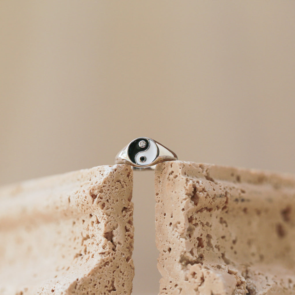 A Yin Yang Ring with a silver enamel finish balances on the edge of a textured beige stone, set against a softly blurred background.