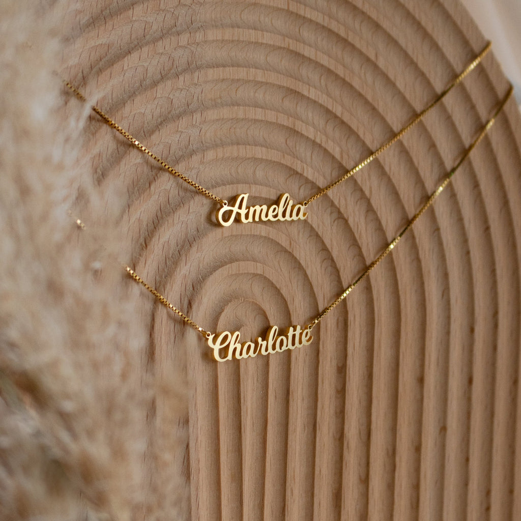Two gold Mellow Double Name Necklaces in Box Chain, customized with "Amelia" and "Charlotte" in a classic font, rest on a textured wooden surface, highlighting elegant personalized jewelry.