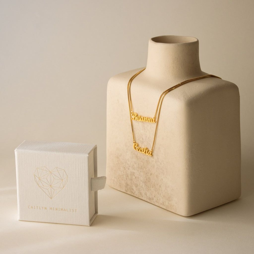 London Double Name Necklace in Box Chain with a chic font on a beige display, next to a white Caitlyn Minimalist jewelry box—perfect personalized jewelry for any occasion.