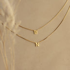 The Alice Double Letter Necklace in Box Chain showcases gold letter pendants B and N on a beige background—a stunning set for personalizing your style.