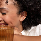 A woman with curly hair smiles, wearing Tiny Pearl Bar Studs and holding a ribbed amber glass—these elegant earrings are perfect for everyday wear.