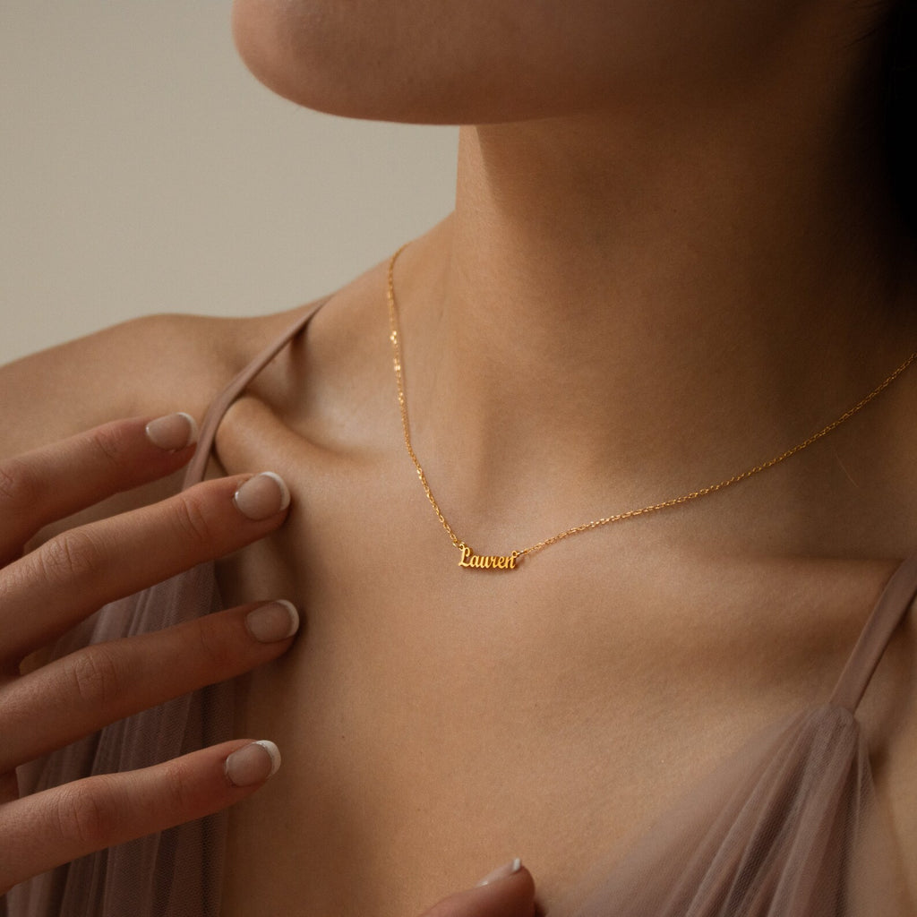 A woman wears the Tiny London Name Necklace in gold with a Lauren nameplate, touching her collarbone with one hand to display its delicate, minimalist style.