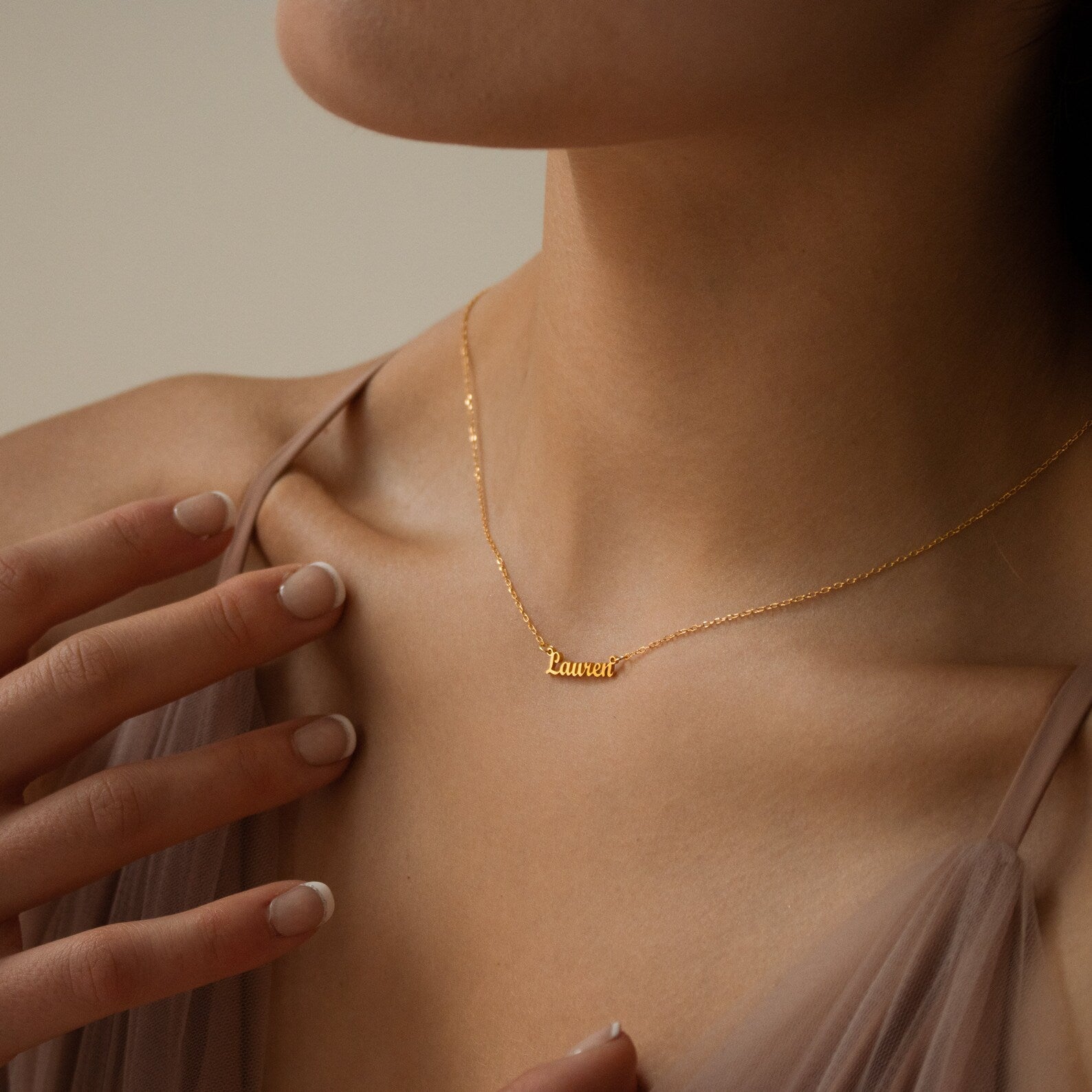 A woman wears the Tiny London Name Necklace in gold with a Lauren nameplate, touching her collarbone with one hand to display its delicate, minimalist style.