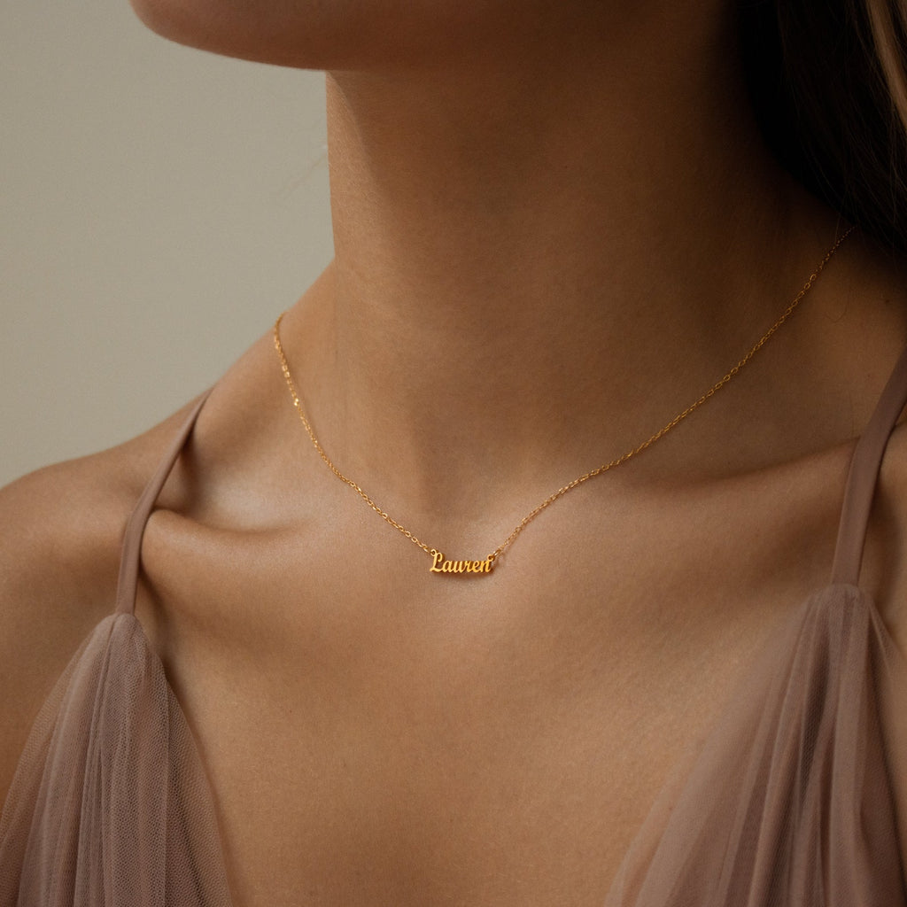 A woman wearing the Tiny London Name Necklace in gold with "Lauren," paired with a sheer taupe top—an ideal minimalist accessory for any occasion.