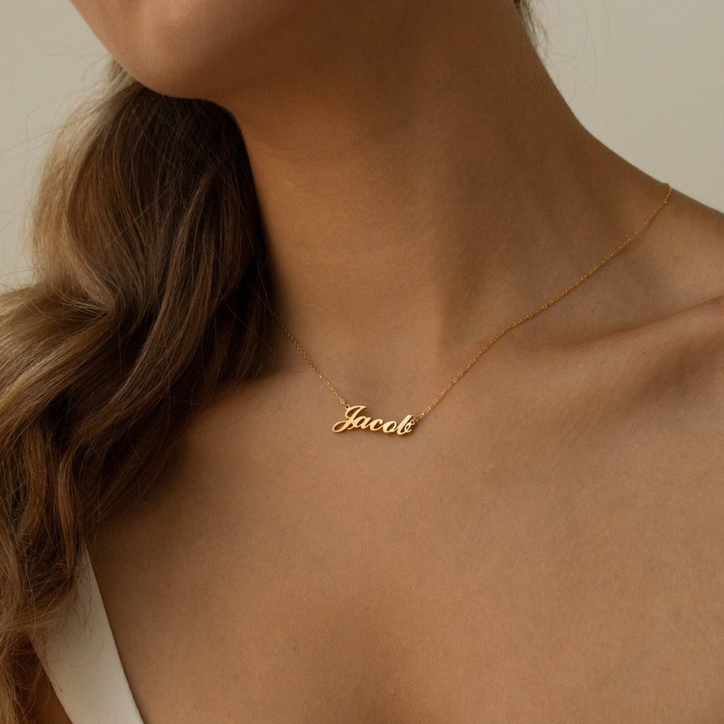 A woman wears the Carrie Name Necklace in gold, showcasing the name Jacob in stylish cursive—a perfect personalized jewelry piece for any look.