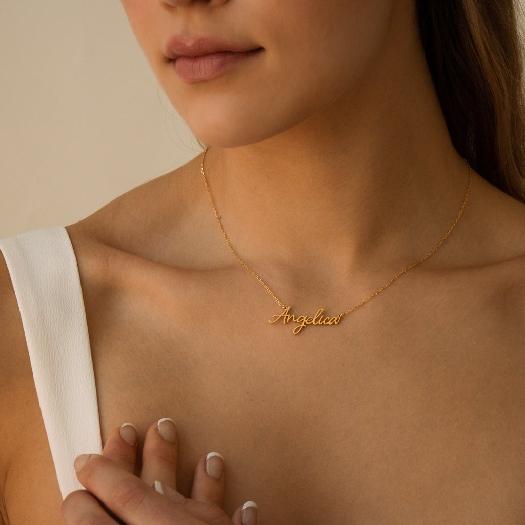 A woman gently touches her collarbone while wearing the Grace Name Necklace, a gold custom piece featuring the name Angelica in elegant cursive script.