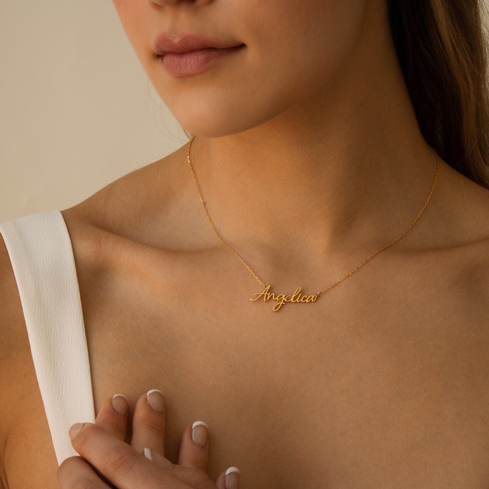 A woman gently touches her collarbone while wearing the Grace Name Necklace, a gold custom piece featuring the name Angelica in elegant cursive script.