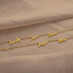 The Nella Multiple Name Necklace in Paperclip Chain featuring gold bold links and custom cursive nameplates—Emily, Jane, Arthur, Jasper, and Elliot—rests on a beige fabric.