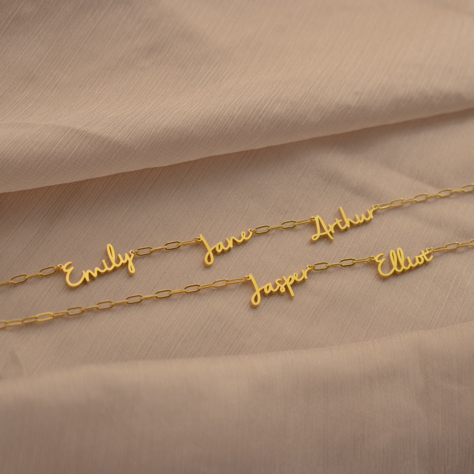 The Nella Multiple Name Necklace in Paperclip Chain featuring gold bold links and custom cursive nameplates—Emily, Jane, Arthur, Jasper, and Elliot—rests on a beige fabric.