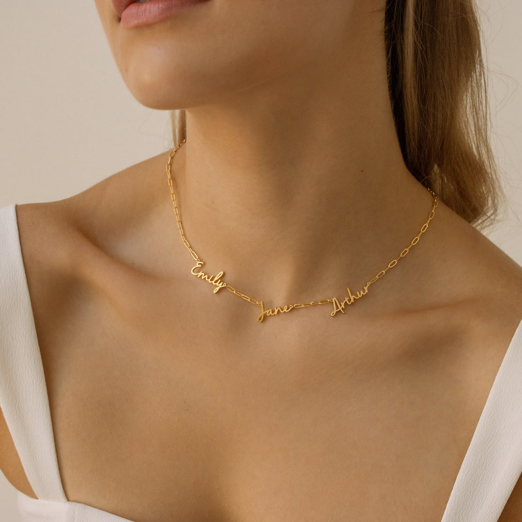 A woman wears the Nella Multiple Name Necklace in Paperclip Chain, displaying Emily, Jane, and Andy in cursive, styled with a white top.