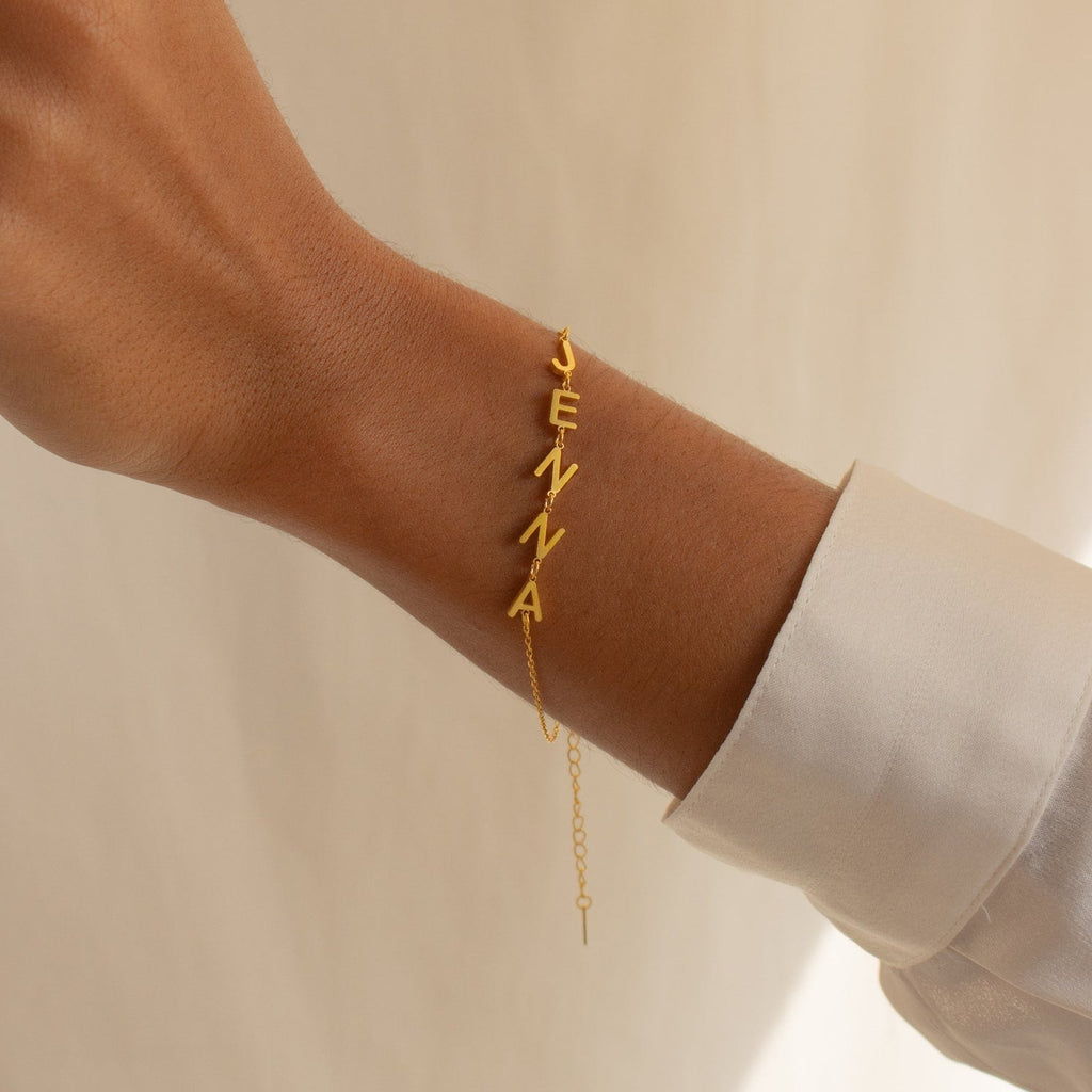 The Custom Letter Bracelet, spelling JENNA in gold, shines on a wrist and pairs perfectly with a crisp white sleeve.