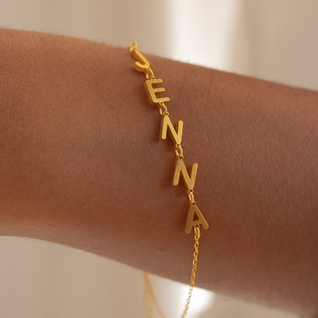 The Custom Letter Bracelet features dangling gold letters that spell JENNA, creating a stunning personalized jewelry piece for your wrist.