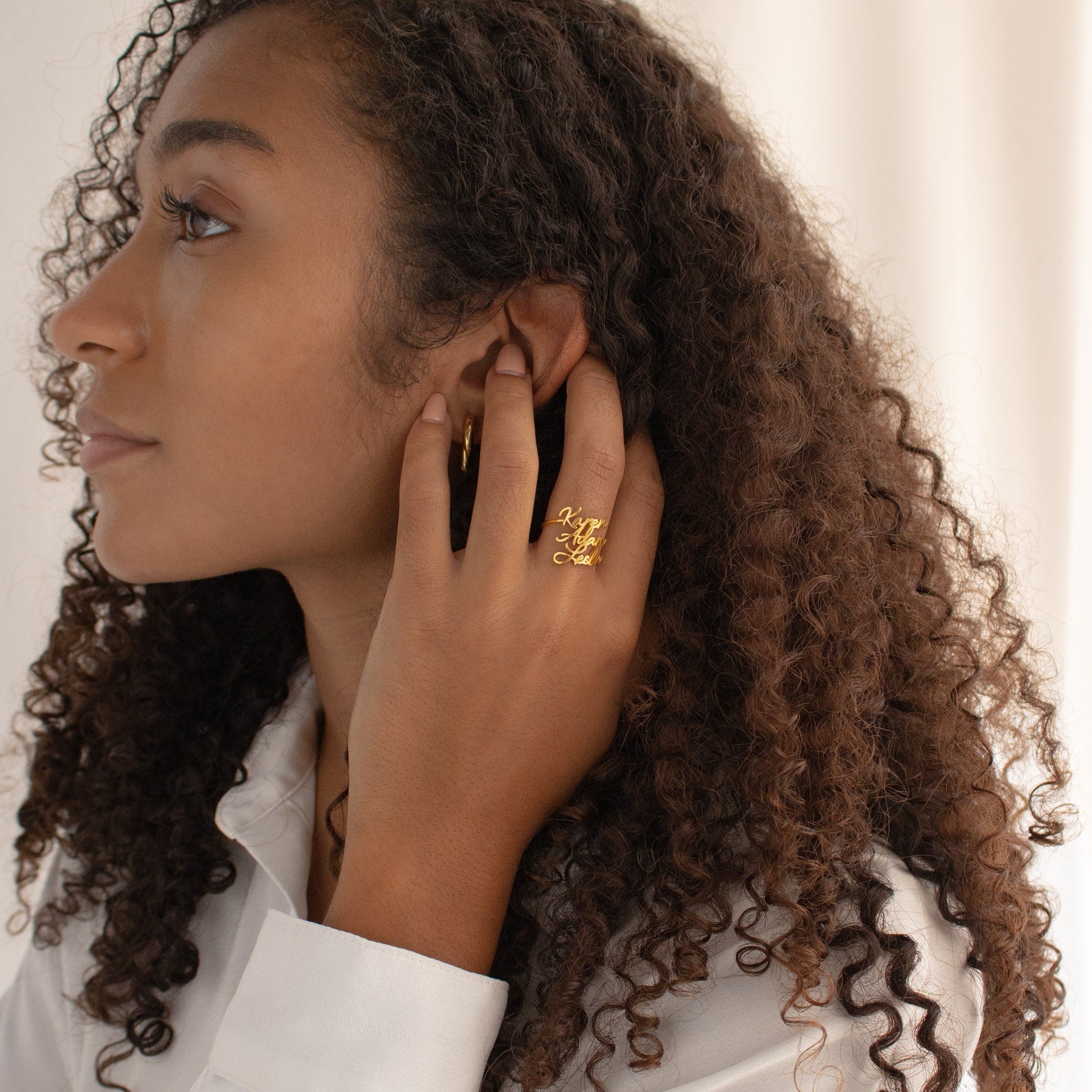 Woman with curly hair wearing the Grace Duo Name Ring and a hoop earring, touching her ear while dressed in a white shirt.