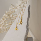 Two Nella Letter Necklaces rest on a textured white stone, framed by dried foliage—perfect for layering or gifting as a personalized accessory.