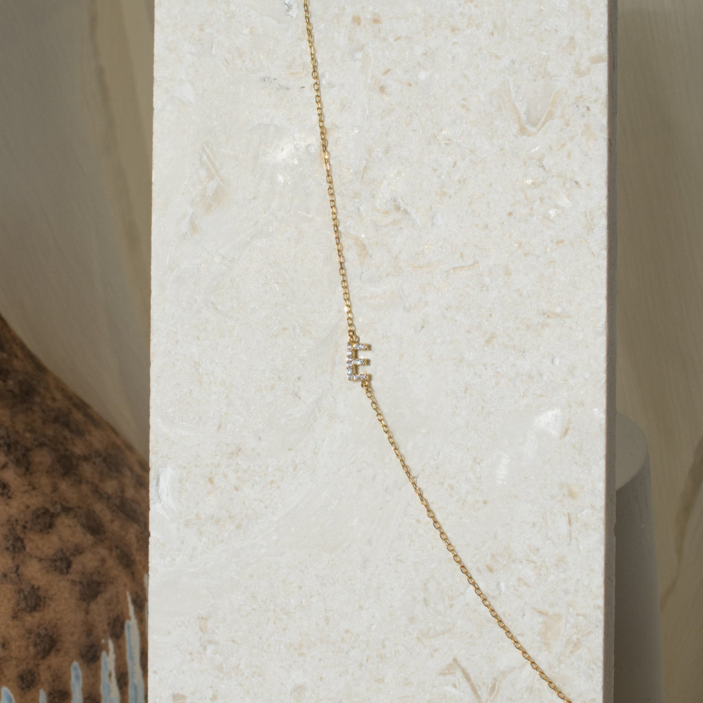 The Pave Sideways Initial Necklace, featuring a gold chain and small vertical rectangular charms, is displayed on a light stone background.