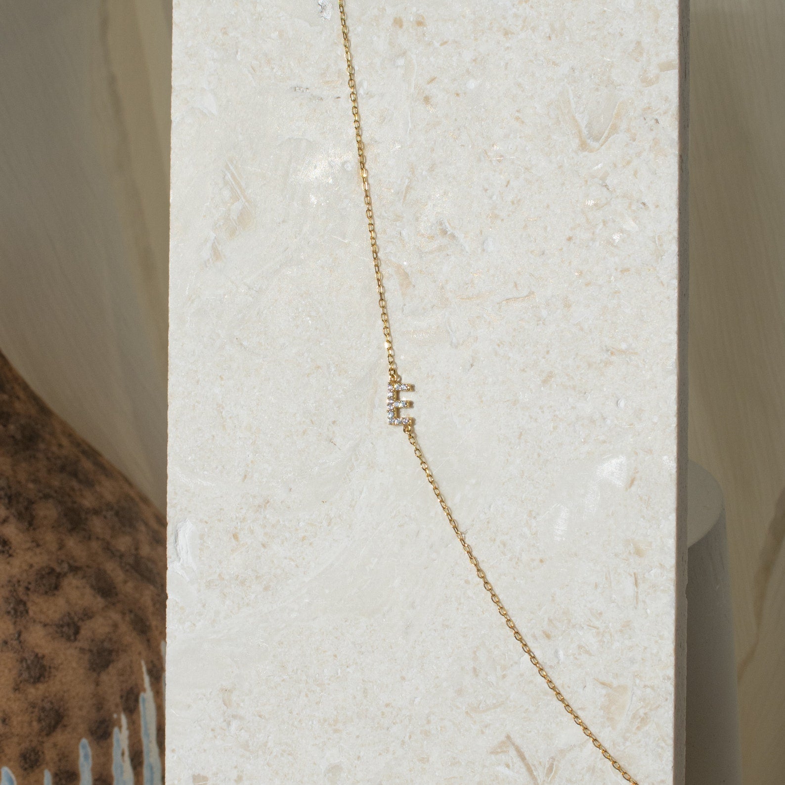 The Pave Sideways Initial Necklace, featuring a gold chain and small vertical rectangular charms, is displayed on a light stone background.