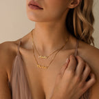 A woman wears the Mellow Double Name Necklace in Box Chain with the names Amelia and Charlotte, paired with a beige dress for a timeless, personalized jewelry look.