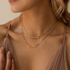 A woman wears the London Double Name Necklace in Box Chain personalized with "Brianna" and "Cecilia," paired with a beige sheer dress, showcasing chic and elegant custom jewelry.