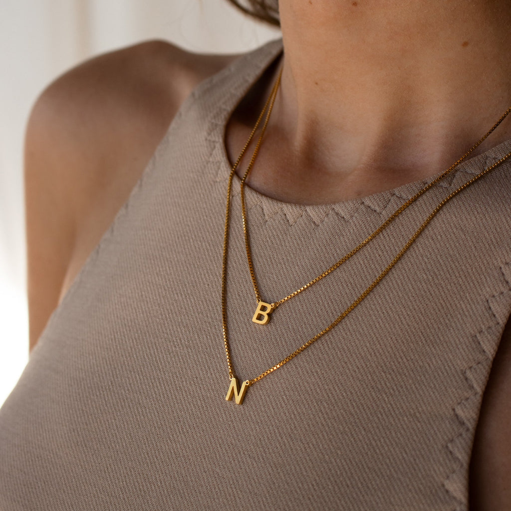 Close-up of a woman wearing the Alice Double Letter Necklace in Box Chain with B and N gold letter pendants over a beige top.