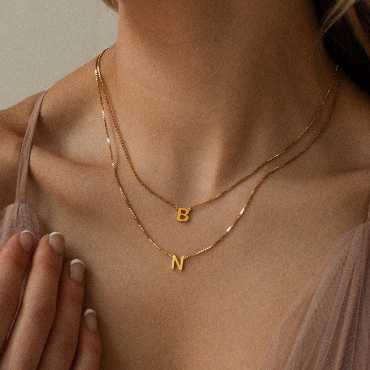 A woman layers the Alice Double Letter Necklace in Box Chain, featuring B and N initial pendants, over a sheer light pink top.